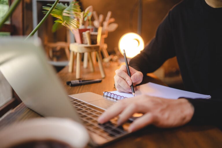 Person writing notes in a notebook while reviewing content on a laptop at a desk