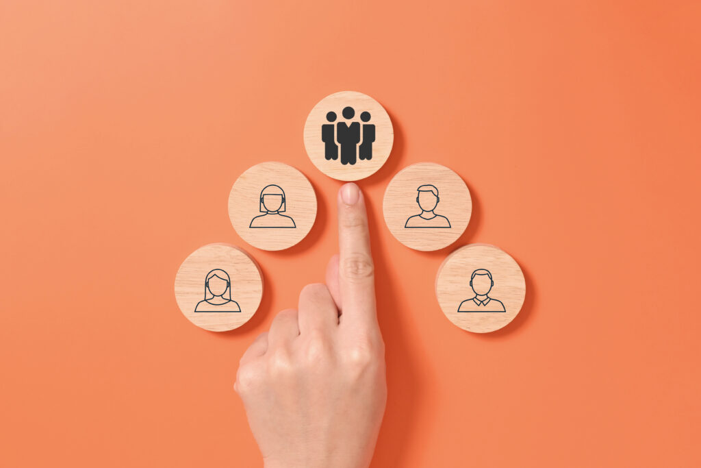 A hand selects a wooden team icon from a group of individual employee icons arranged on an orange background.